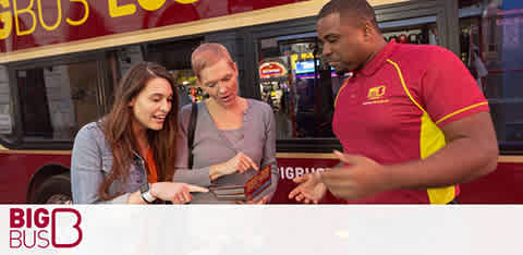 Description: The image features three individuals interacting in front of a 'Big Bus' tour bus, which is partially visible in the background. On the left, a young woman with long hair is smiling and looking at a brochure she is holding. Next to her, an older woman, possibly in her 60s, with short hair, leans in to also view the brochure with an interested expression. They are both examining the brochure together. On the right, a man dressed in a Big Bus uniform, which includes a red shirt with yellow and white trim, is animatedly gesturing and appears to be explaining something to the two women, possibly about the bus tour. He is looking at them and speaking as they listen. The Big Bus logo is prominently displayed in the top left corner of the photo, signaling the brand of the tour service. Catch the excitement of exploring the city and don't miss out on the savings — GreatWorkPerks.com offers the lowest prices on tickets for your next adventure!