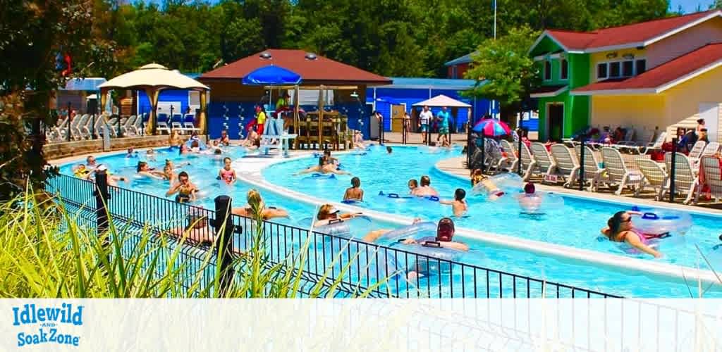 Image shows a lively outdoor water park on a sunny day. Visitors enjoy floating in a lazy river, lounging on deck chairs, and relaxing under blue parasols. Brightly colored buildings and lush green trees frame the scene, creating a cheerful, family-friendly atmosphere. The words 'Idlewild SoakZone' are visible, indicating the park's name.