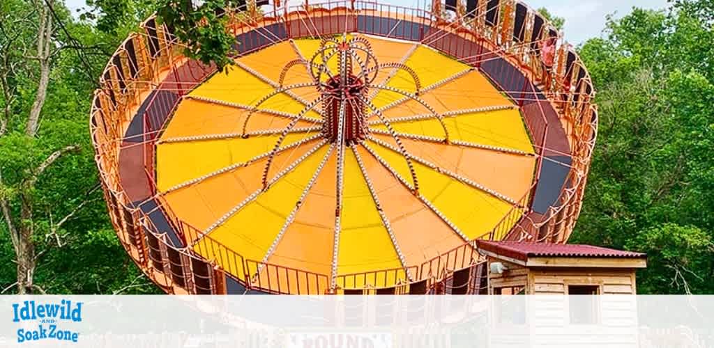 Image shows a large, stationary Ferris wheel with vibrant orange and yellow coloring, set against a backdrop of green foliage. The structure is adorned with intricate patterns and positioned behind a small building with a brown roof. The Idlewild & SoakZone logo is visible in the bottom left corner.