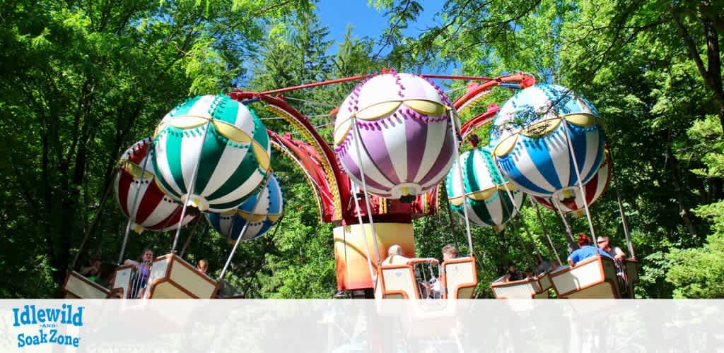 Image features a colorful balloon ride at Idlewild SoakZone. Sunlit trees surround the ride where multiple striped balloon-shaped cars with baskets carry smiling riders through the air. The backdrop is a clear blue sky on a bright day. The ride evokes a sense of fun and adventure in a forest setting.