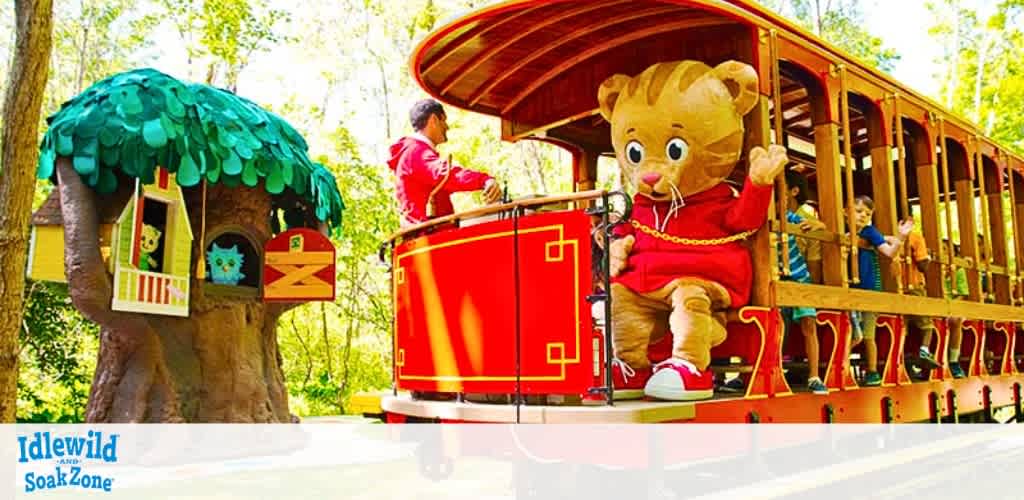 A colorful open-air train with a red and yellow exterior glides through a green, forested area. A costume character resembling a large, friendly lion in red attire waves from the rear of the train. Children and a train operator are seen enjoying the ride, with whimsical structures like a tree with a face and mailbox nearby, all under a bright, sunny sky. The text Idlewild and SoakZone is displayed at the bottom.