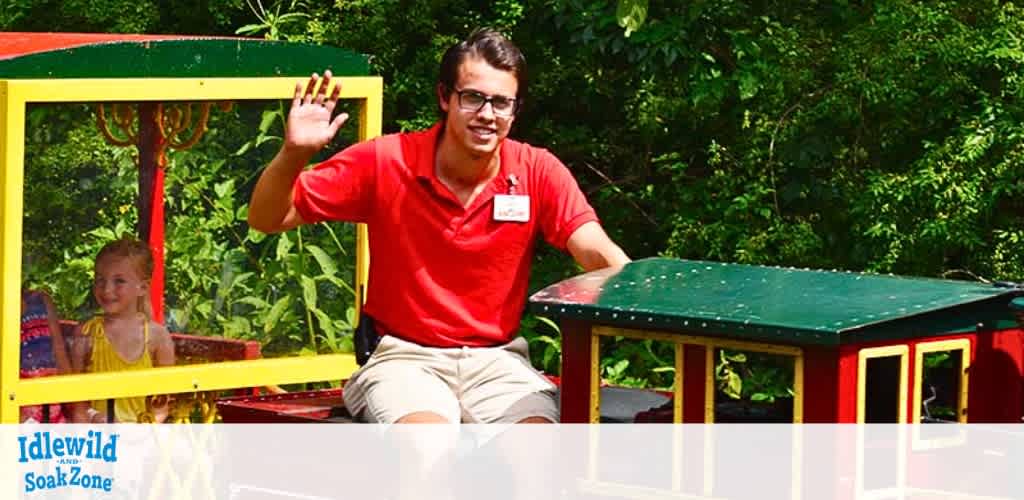 Image of a smiling train attendant in a red shirt waving with one hand, seated on a colorful children's train ride at Idlewild SoakZone. In the background, a delighted child peeks from a yellow-framed window, adding to the cheerful, vibrant scene at the amusement park.