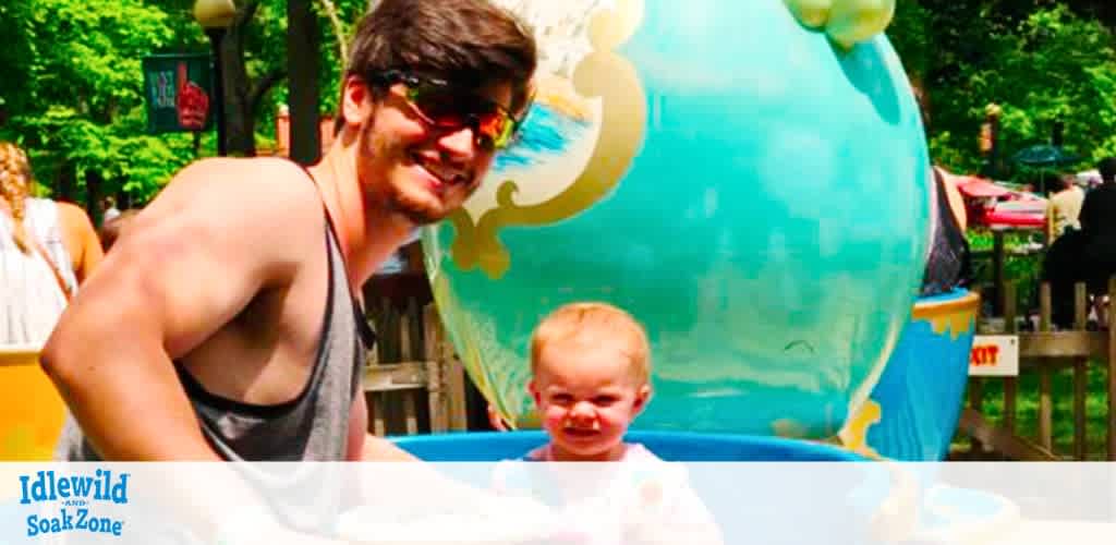 A man in a tank top and sunglasses smiles as he enjoys time at a water park. Next to him, a young child gleefully peeks out from behind a blue water play structure. The background is vibrant with greenery, and a globe-like decoration adds to the fun atmosphere of Idlewild SoakZone.