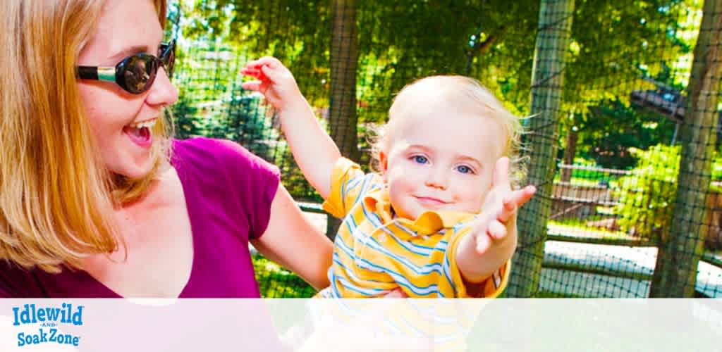 A smiling woman in sunglasses and a purple top is playfully interacting with a cheerful toddler in a striped yellow shirt. They are outside with greenery in the background, and the words 'Idlewild & SoakZone' appear, suggesting they are at a park. The child seems to be reaching out towards the camera with a joyful expression.