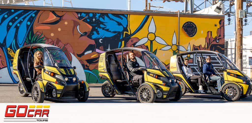 Image shows three yellow and black GoCar tour vehicles lined up with drivers seated, in front of a colorful mural with a nature theme. The mural depicts a lion and abstract designs with a blue and red color palette. The GoCar logo is visible at the bottom of the image.