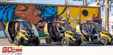 Image shows three small, yellow and black GoCar vehicles in a row with drivers seated inside. They are parked in front of a vibrant, colorful mural depicting abstract and floral designs with a sun motif. The GoCar logo is visible at the bottom. The setting appears to be an urban environment with clear blue skies.