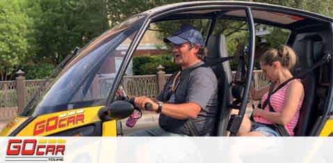 Image shows two people in a small yellow and black open-topped vehicle, branded 'GoCar'. A man in the driver's seat is smiling and holding the steering wheel, while a woman on the passenger side looks down, possibly at a map. Trees and a park setting are visible in the background. The scene suggests a leisurely urban tour.