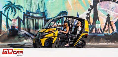 Image of two smiling individuals enjoying a ride in a yellow and black GoCar in front of a vibrant mural featuring palm trees and landmarks. The GoCar logo is visible at the bottom. The scene suggests a fun urban adventure.