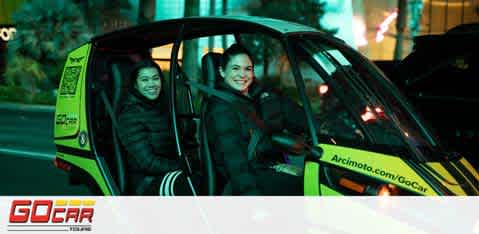 Image shows two people smiling inside a compact, yellow and black vehicle with a clear door. They are wearing seatbelts and appear to be enjoying a night drive. Visible logos suggest it's a GoCar tour service. The scene is illuminated by streetlights and hints at an urban setting.