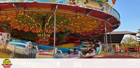 Image of a colorful carousel at an amusement park with bright lights and decorative panels, featuring various carousel horses and seating areas. The ride is surrounded by safety railings, and a clear blue sky can be seen overhead. The corner features a logo that reads 'FUN SPOT.'