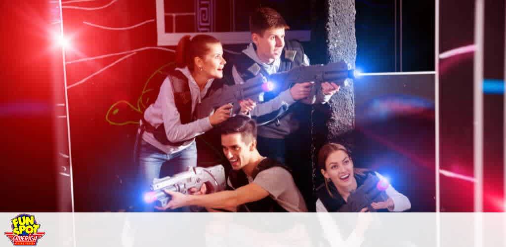 Image shows four people enjoying a game of laser tag, with two individuals in the foreground poised with laser guns, one crouching and one standing. Two more participants are seen in the background, using a wall as cover. They are all wearing dark vests and aiming their laser guns enthusiastically. The environment is dimly lit with neon lights enhancing the action-packed atmosphere. The Fun Spot America logo is visible in the lower left corner.