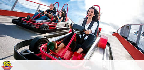 The image shows a group of enthusiastic individuals racing in go-karts on an outdoor track. The person in the forefront is a woman with her hair swept back by the wind, wearing a black and white outfit, expressing joy as she steers her kart with focus. A clear sky is above and the track stretches ahead with vibrant colors. The logo for Fun Spot appears in the corner, adding to the lively atmosphere.