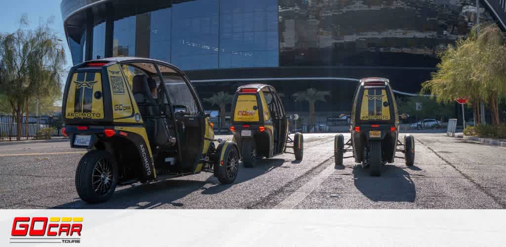 This image features three yellow and black, three-wheeled GoCar tour vehicles parked on a concrete roadway. Each vehicle is designed with a single rear wheel and two front wheels, with a clear, open cabin structure for driver and passenger seating. The backdrop reveals a modern building with curved architecture and reflective glass surfaces. A bright, clear sky suggests a sunny day with several palm trees flanking the left side, providing a bit of greenery to the urban environment.

At FunEx.com, we are committed to offering exciting experiences, and the thrill of exploring in a GoCar is no exception. Don't forget to check out our exclusive discounts for the lowest prices on tickets to the best attractions and tours.