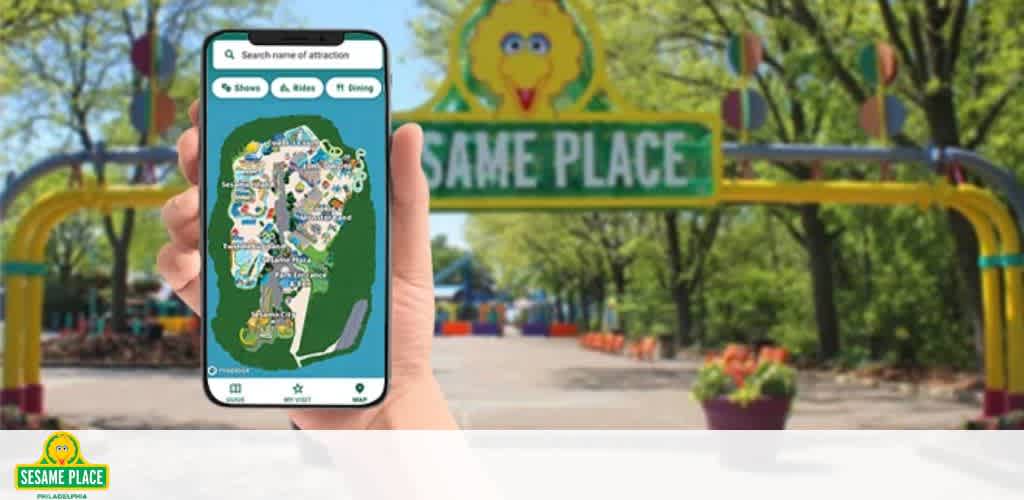 Foreground: A person's hands holding a smartphone with a park map on the screen. Background: Entry gate of Sesame Place with signage and Big Bird's image. The atmosphere is bright and inviting. Flowers in bloom are visible.