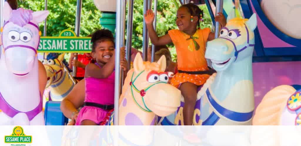 Two joyful children are having fun on a carousel at Sesame Place. One child in a pink dress, another in orange attire, both with arms raised in excitement, riding whimsical horses. A bright, colorful Sesame Street sign adds to the cheerful atmosphere.