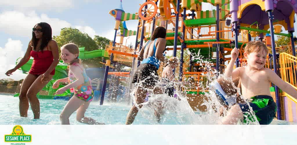 Image shows happy children and adults at a Sesame Place water park. They are splashing in the shallow water with bright, colorful water play structures in the background under a sunny sky. The Sesame Place logo is visible in the corner.