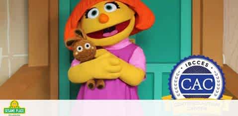 Image features a smiling orange-haired puppet from Sesame Place wearing a pink dress and holding a brown toy bear. A blue seal with text indicates certification by the International Board of Credentialing and Continuing Education Standards. The background is simple with light colors.