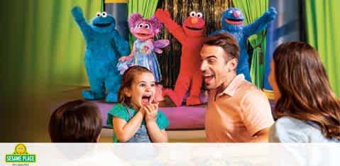 A joyful family moment with two adults and a child excitedly watching a stage show featuring three colorful Sesame Street characters. The child appears delighted with hands on cheeks, and the adults share the child's excitement, overlooking a brightly lit performance area with vibrant green curtains. The Sesame Place logo is visible in the corner, suggesting a themed entertainment experience.