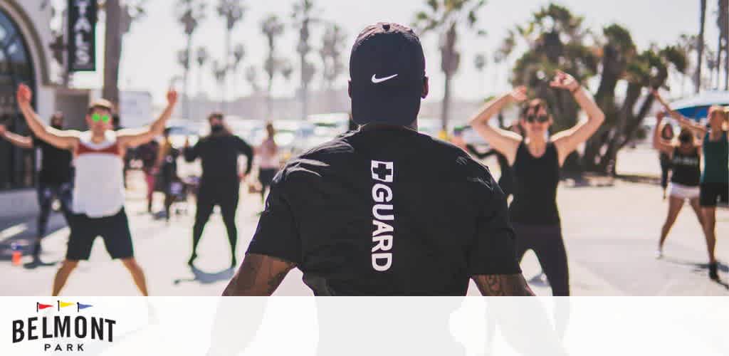 A lifeguard watches over a lively outdoor fitness class at Belmont Park. Sunlit participants, some in motion, enjoy the energetic atmosphere under a clear sky, with palm trees lining the background, creating a scene of active community and vigilant supervision.