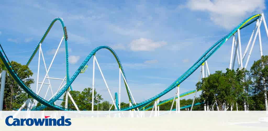 Image shows a roller coaster at Carowinds theme park with a clear blue sky. The coaster's track is teal with white supports, featuring a series of high peaks and steep drops among a backdrop of lush green trees.