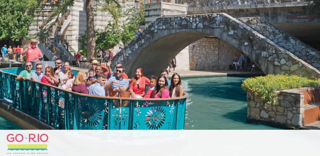 Image description: A group of smiling people enjoying a sunny day on a boat tour along a peaceful river. They are passing under a stone arch bridge adorned with greenery. The blue railing of the boat features decorative cut-outs of sunbursts. Text on the image reads 'GO RIO San Antonio River Cruise'.