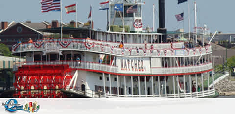 Image of a large, traditional paddle-wheel riverboat with multiple decks, a white superstructure with red accents, and a red paddle wheel at the rear. Flags are flying atop. The boat is docked with a cityscape in the distance.