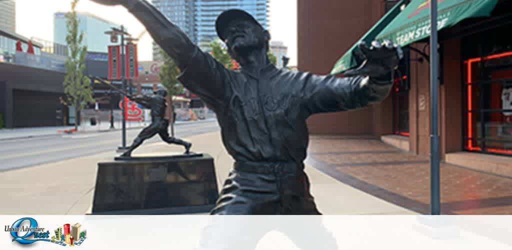 Image of a statue of a baseball player mid-pitch, with arm extended in front of a city street. Another similar statue is in the background. The statues are positioned on pedestals outside a building with a green awning. Traffic signs and urban architecture are visible. The Ultimate Adventure Quest logo is at the bottom left.