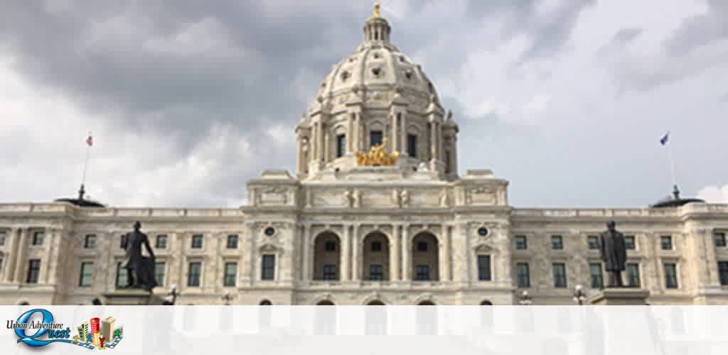 Image displays a grand, neoclassical building with a prominent central dome and multiple levels of intricate stonework. The dome is topped with gold ornamentation. Flanking the central structure are symmetrical wings with rows of windows and flags flying atop. A statue is visible in the foreground, and the sky behind is partly cloudy. The image features a logo in the bottom left corner suggesting a connection to an adventure or event company.