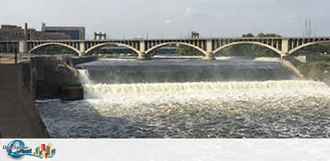Image of a multi-arch bridge spanning a river, with water cascading over a dam in the foreground. The sky is clear, and buildings can be seen in the background. A logo is visible at the bottom right corner.