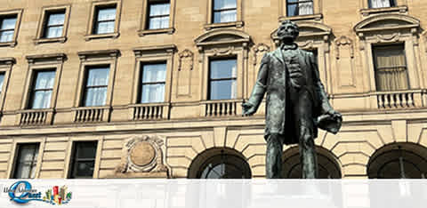 The image shows a full-length statue of a historical figure standing before a classic building with multiple windows and ornate detailing. The figure is dressed in a coat and trousers, possibly from the 19th century, holding what appears to be a hat at his side. The façade of the building has decorative elements above the first-floor level. Skies are clear above the scene.