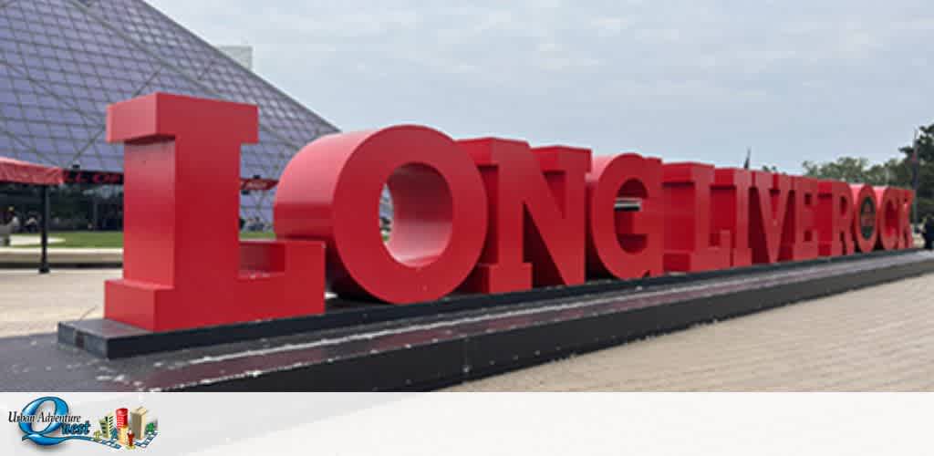 Image displays large red three-dimensional text spelling  LONG LIVE ROCK  set against an outdoor backdrop with pyramid-shaped glass building in the distance. There's a clear sky above and paved ground below the bold structure.