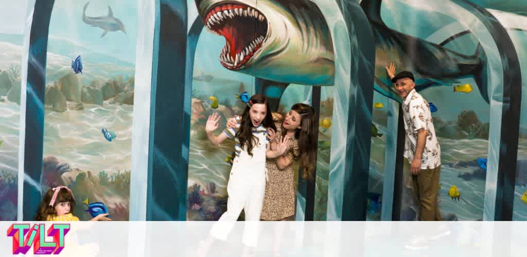 Image depicts three individuals interacting with a 3D ocean mural that creates an illusion of them being underwater. The artwork features a large open-mouthed shark and various marine life. A child on the left appears to be peering out from an underwater cave, a young woman and another individual in the center seem to be playfully reacting to the shark, and a man on the right is positioned as if he is inside a bubble column, smiling and touching the mural.