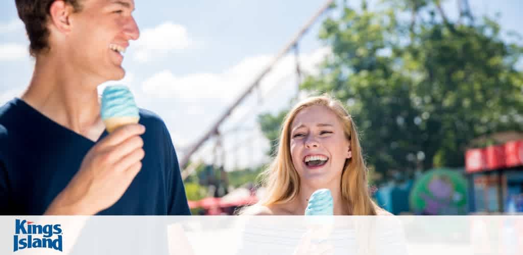 A joyful scene at Kings Island amusement park with a young man in a navy shirt enjoying a blue ice cream cone and a smiling young woman with blonde hair, sharing a light-hearted moment under a clear sky. Faint outlines of park rides and greenery are in the background.