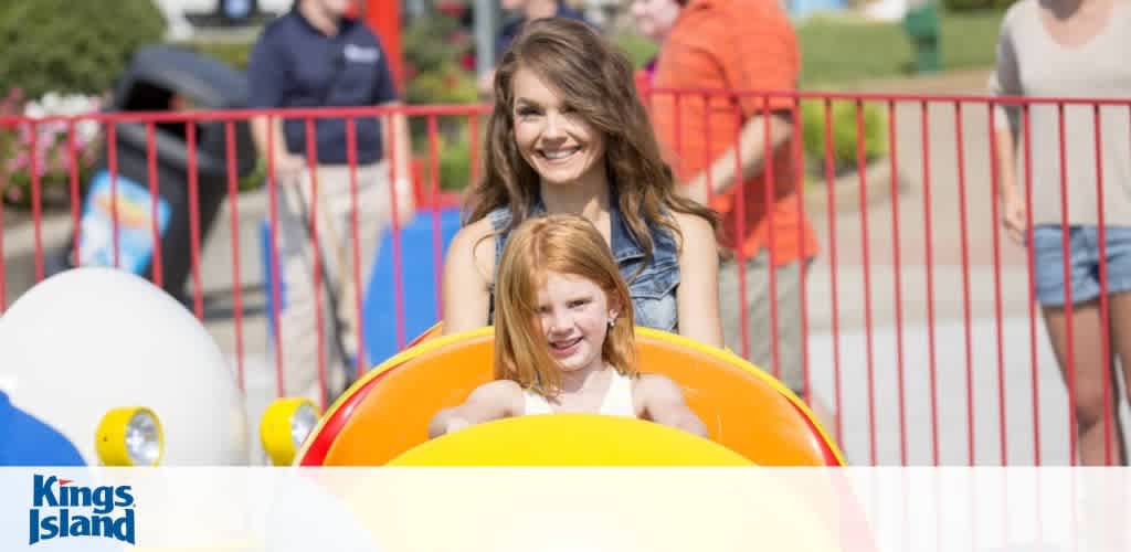Image shows a joyful young woman and a little girl with red hair riding in a yellow amusement park car. They are smiling and appear to be enjoying a sunny day out. The background includes other guests and park structures, with a Kings Island logo visible.