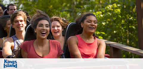 Group of people expressing excitement on a roller coaster ride at Kings Island. They are seated in a row with expressions of joy and anticipation. Trees in the background suggest an outdoor setting. The atmosphere is energetic and fun.