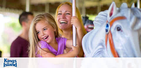 A young girl with blonde hair, wearing a lavender top, and a woman, likely her mother, with brown hair, wearing a white top, are joyfully riding a carousel. The girl is in front of the woman, both holding onto the carousel pole, smiling brightly. A whimsical carousel horse is partially visible on the right. They appear to be having a delightful time at Kings Island amusement park, as indicated by the logo in the corner.