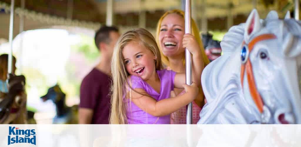 A joyful young girl with light hair, wearing a purple top, and a smiling woman with blonde hair in a pink tank top are enjoying a carousel ride. In the foreground, a white carousel horse with a blue eye and red bridle accents adds a playful touch. The background is softly focused with the Kings Island logo at the bottom.