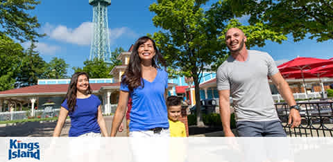 A cheerful family with two adults and two children are walking with smiles on their faces at an amusement park on a sunny day. Trees, a tall tower structure, and park buildings can be seen in the background. The park's logo, Kings Island, is visible in the photo's lower-right corner.