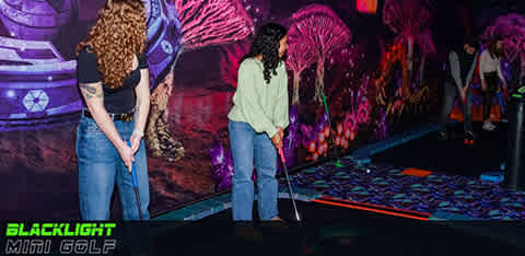 Two people are playing mini golf in an indoor, blacklight-lit course with vibrant neon wall murals. The person in the foreground is preparing to take a shot while the other watches. The environment gives off a fun and unique glow, adding to the entertainment experience. A text overlay says 'Blacklight Mini Golf'.