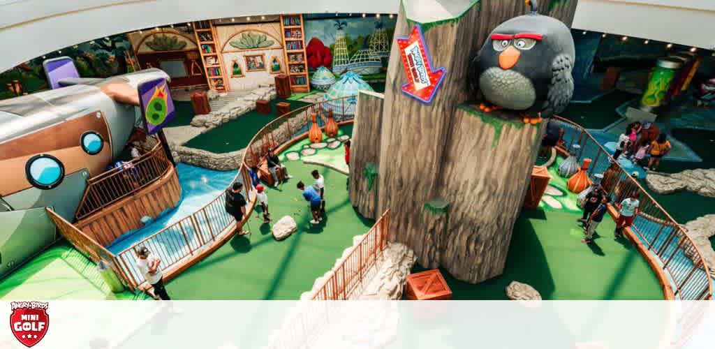 The image showcases an indoor mini-golf course with an Angry Birds theme. On the left, people are playing near a wooden, propeller-driven airplane. A large, animated grey bird statue watches over the course on the right, where more individuals enjoy golfing amongst vibrant game-inspired decorations.