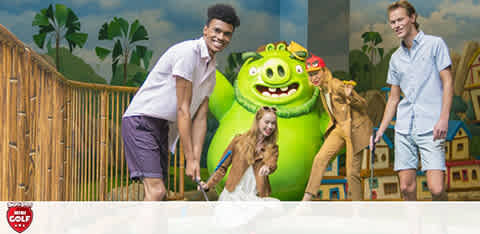 Three individuals enjoy mini-golf beside a bright green, friendly-looking inflatable character. They exhibit cheerful expressions matched by a whimsical tropical backdrop, complete with cartoon-like palm trees and colorful houses, creating a vibrant, playful atmosphere typical of casual outdoor activities.