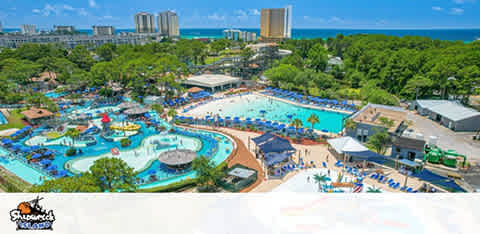 Aerial view of a waterpark with pools and slides, surrounded by trees and high-rise buildings in the background.