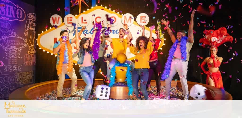 A vibrant group of people celebrates with excitement on a colorful casino-themed stage. They throw confetti into the air, surrounded by large dice, casino decorations, and bright lights. A joyous atmosphere conveys a fun-filled event, complete with a backdrop that reads  WELCOME  in bold letters, capturing the essence of party and play at Madame Tussauds.