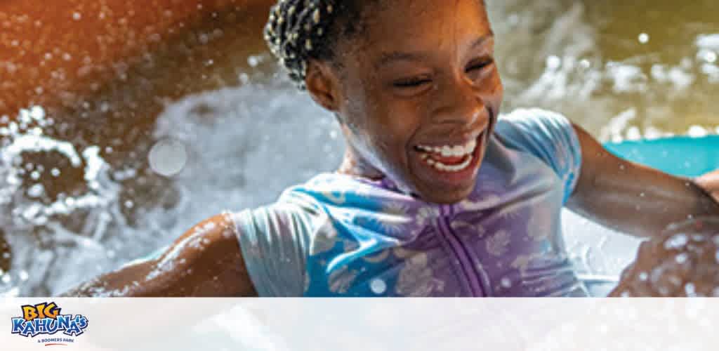 Image displays a young girl with a joyous expression playing in water. She is wearing a blue swim shirt and the sun highlights the water droplets around her. The logo for Big Kahuna's, a water park, is visible in the top left corner, suggesting a fun-filled environment for family activities.