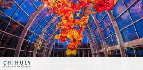 Image shows the interior of a glasshouse with a high arched ceiling. Numerous bright, fiery-hued glass sculptures hang from the ceiling, resembling flowers or sea creatures. The sky outside is twilight blue. Below the image, the words 'Chihuly Garden and Glass' are visible.
