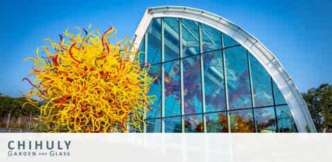 Image features a vibrant yellow and orange glass sculpture resembling a burst of twisting tentacles in front of a modern curved glasshouse under a clear blue sky. The words Chihuly Garden and Glass are displayed at the bottom center.