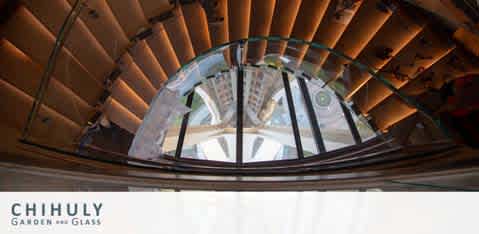 Interior view looking up at a curved glass ceiling with a large, circular skylight at the center. Spiral wooden staircases frame the sides, leading the eye to the skylight above. The white text at the bottom reads 'Chihuly Garden and Glass.' The setting is indoors with natural light filtering through the glass dome.