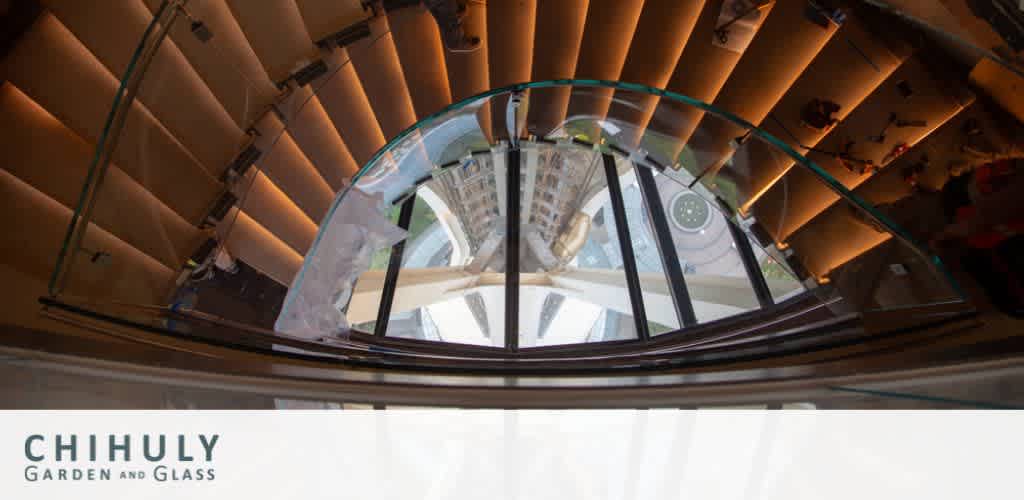 Image of a spiral staircase viewed from below, showcasing the geometric patterns of the steps and the curved glass railing. The daylight filters in from the circular glass ceiling above. Visible in the corner is the Chihuly Garden and Glass exhibit logo.