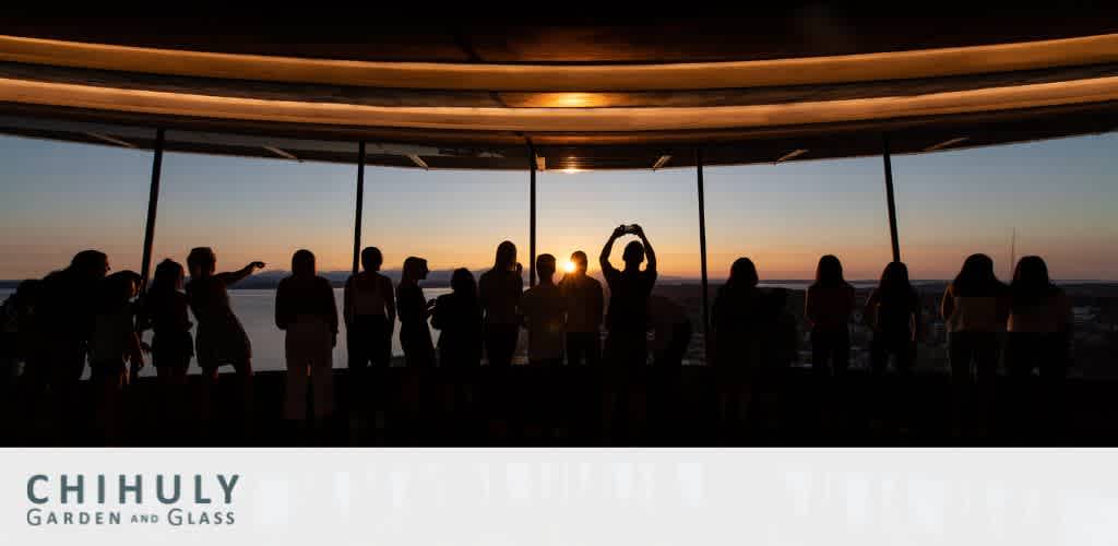 An observation deck at dusk with silhouettes of people admiring the view. One individual at the center forms a heart shape with their hands towards the sky. The Chihuly Garden and Glass logo is visible at the bottom. Warm light from the setting sun enhances the tranquil ambiance.