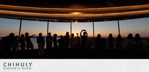 Silhouettes of visitors at a viewing deck with a sunset background. The sky transitions from warm orange to deep blue. In the foreground, a circle is formed by a person's arms, framing the sun. Underneath, text reads 'CHIHULY GARDEN AND GLASS'.